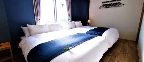 Una cama bien hecha con ropa de cama blanca y azul, una ventana con cortinas, un aire acondicionado de pared y un perchero con ganchos.