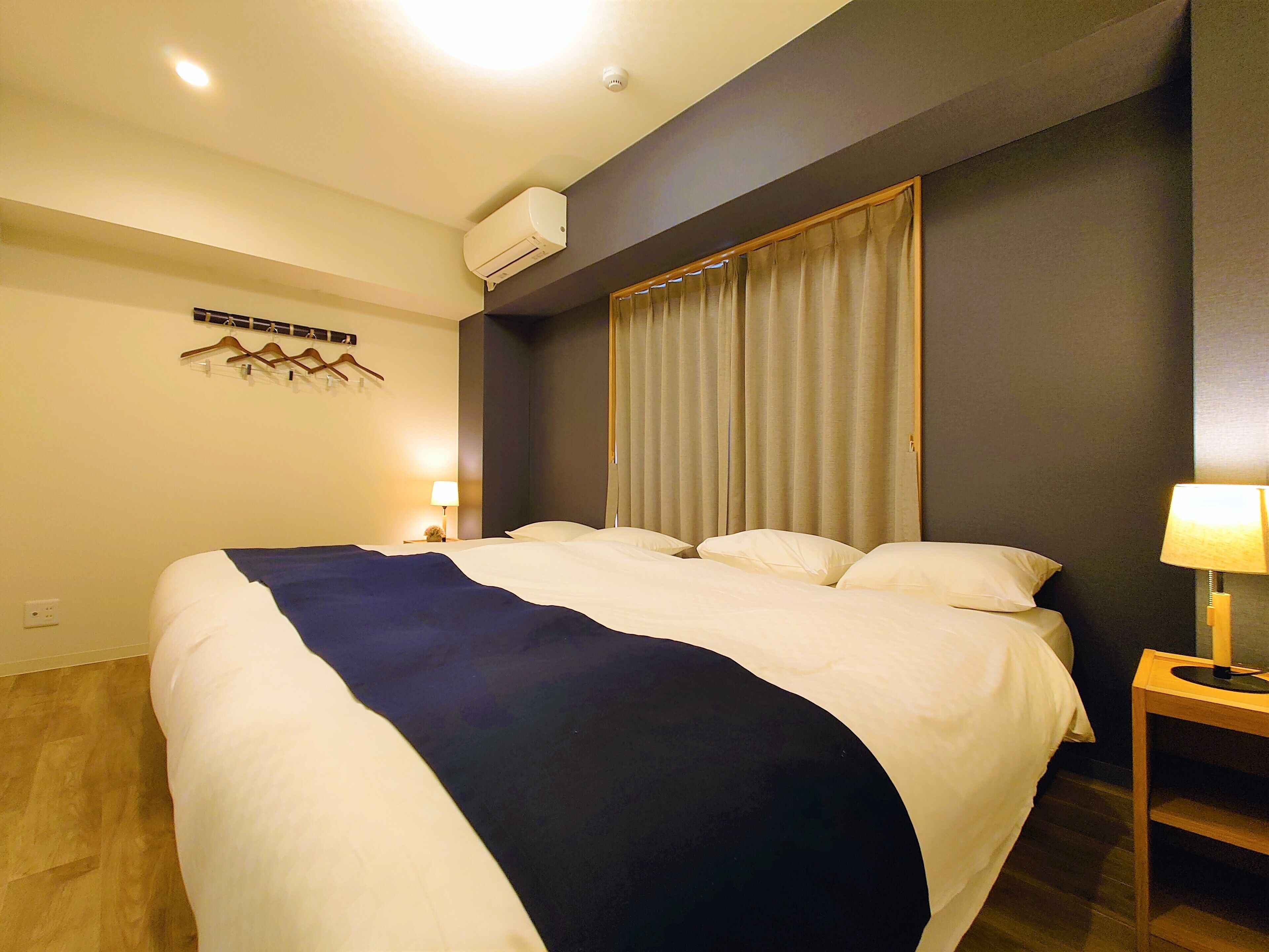 A hotel room with a large bed, a bedside table with a lamp, a wall-mounted air conditioner, and a curtain.