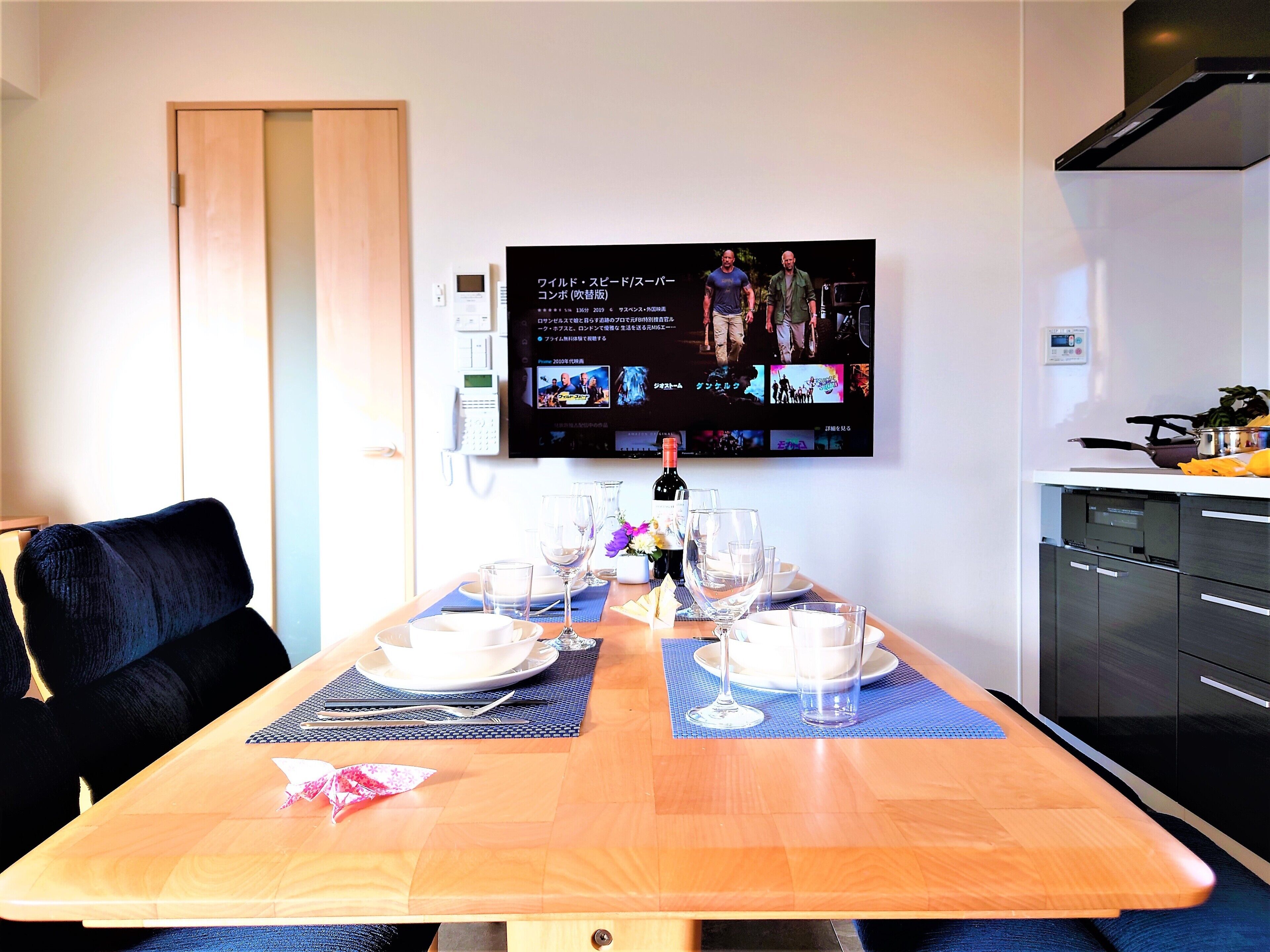Un comedor con una mesa de madera dispuesta para cuatro personas, un televisor montado en la pared mostrando una película y una cocina con electrodomésticos modernos.