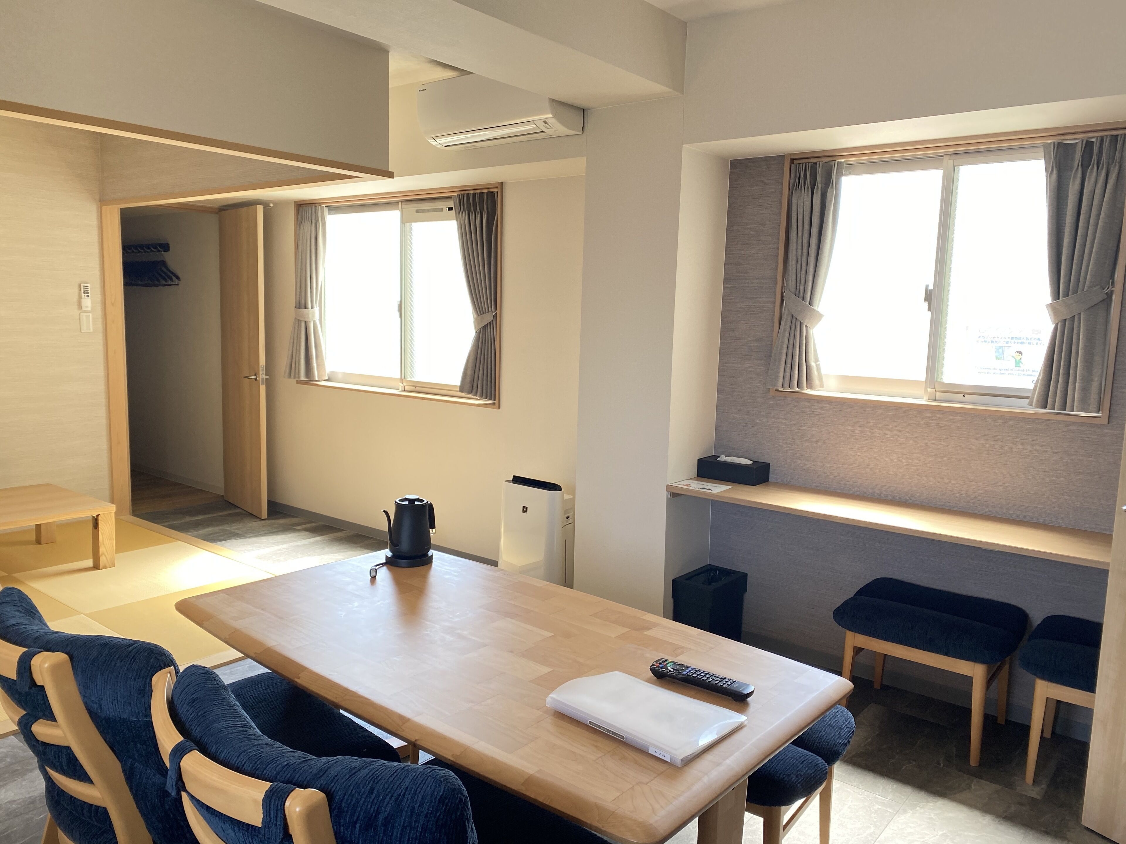 Ein modernes, kompaktes Zimmer mit einem Holztisch, zwei blau gepolsterten Stühlen, einem kleinen schwarzen Wasserkocher, einer Fernbedienung und einem weißen Laptop auf dem Tisch. Zwei Fenster mit Vorhängen, eine eingebaute Bank und eine kleine Ablage mit einer Taschentuchbox sind ebenfalls vorhanden.