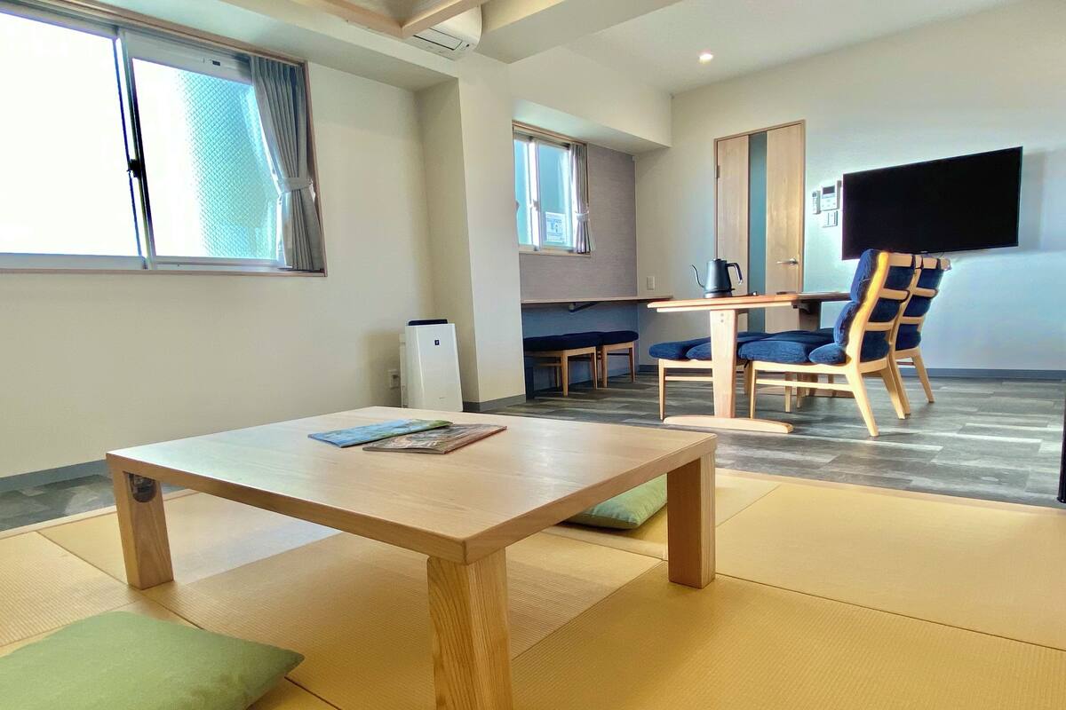 Una habitación con esteras de tatami, una mesa de madera con libros, un televisor y una zona de kitchenette.