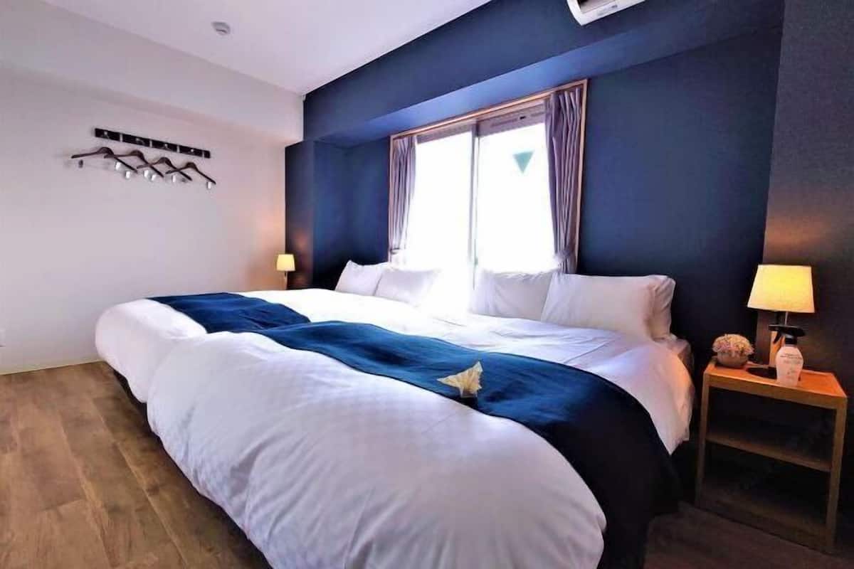 Una habitación de hotel con una cama grande, una mesita de noche con lámpara, una ventana con cortinas y un perchero en la pared.