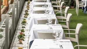 A row of tables with white tablecloths and wicker chairs, set against a backdrop of a green leafy wall and a row of cacti. - Hotel Villa Pandora (Maiori)