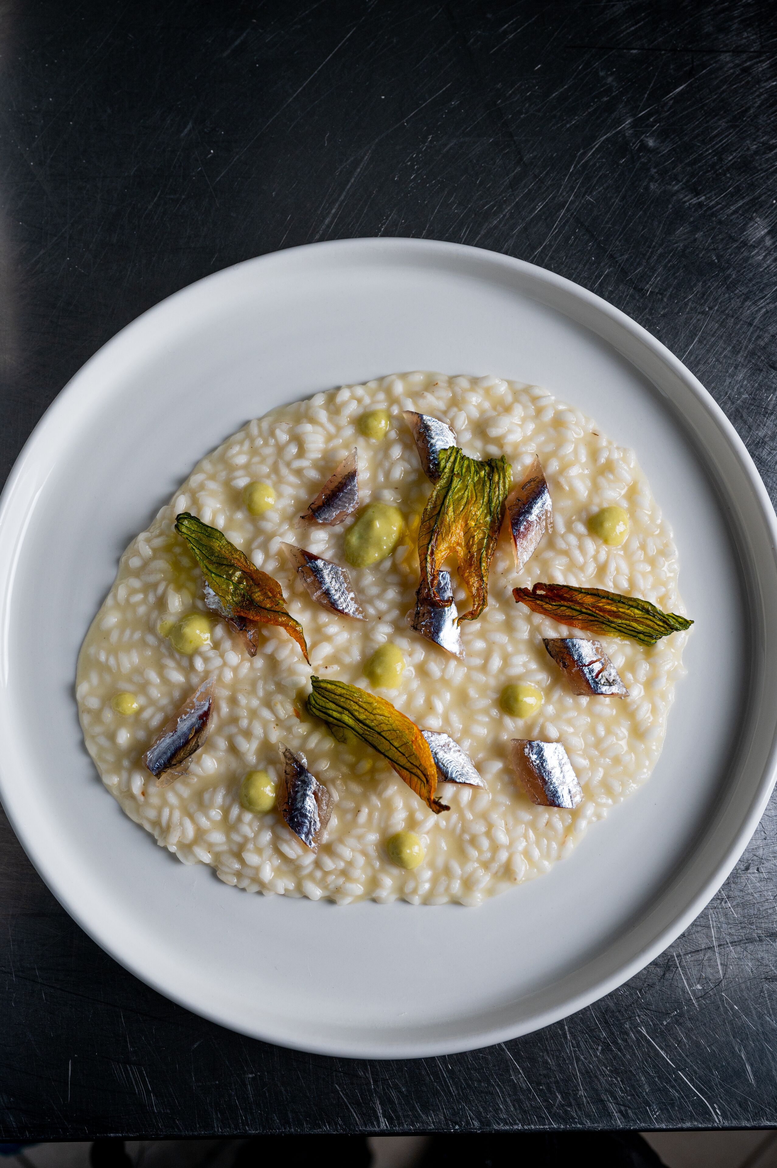 Ein weißer Teller Risotto mit grünen Pistazien und frittiertem Fisch auf dunklem Untergrund.