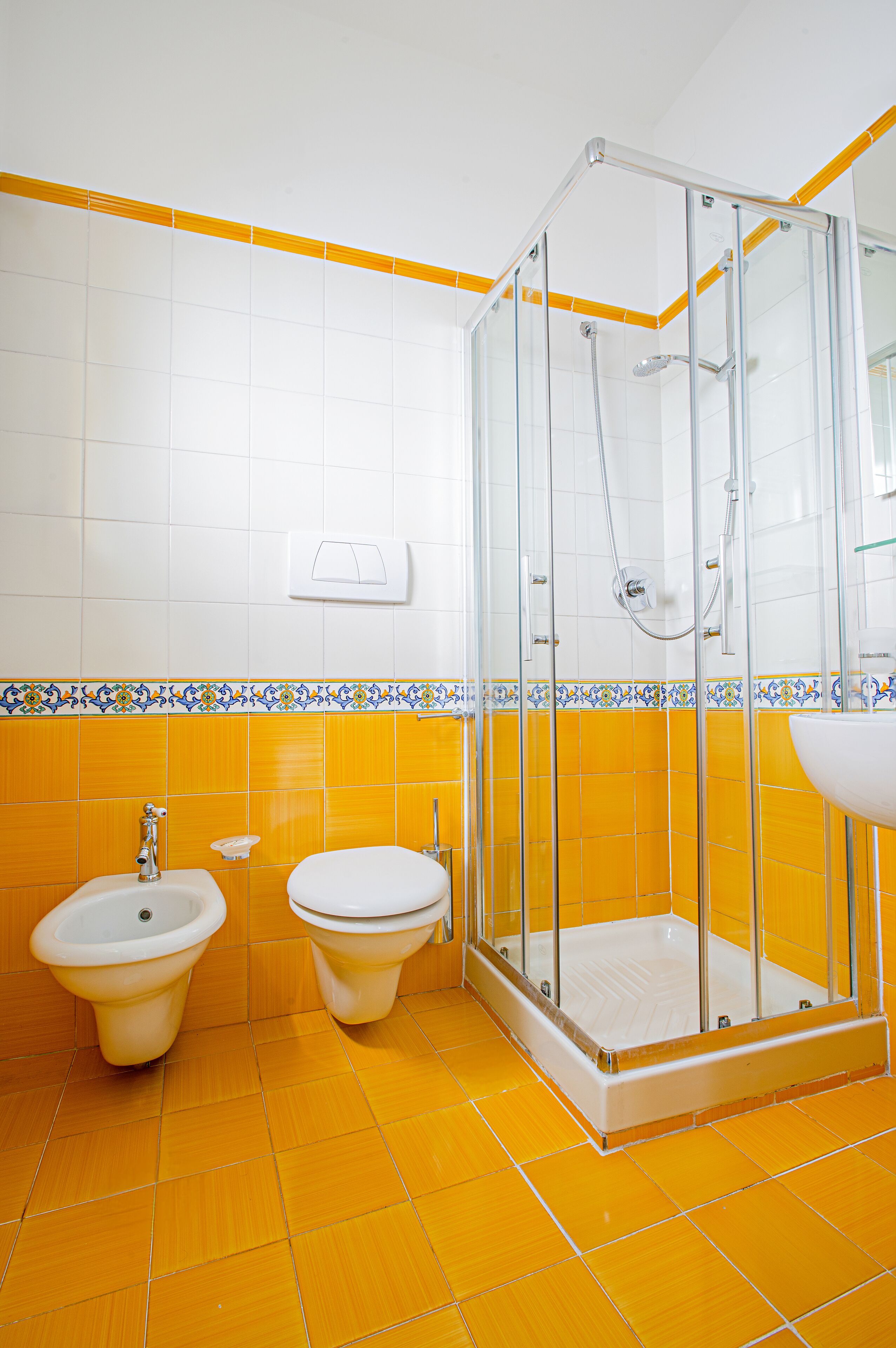 A bathroom with yellow tiles, a glass shower enclosure, a sink, and two toilets.