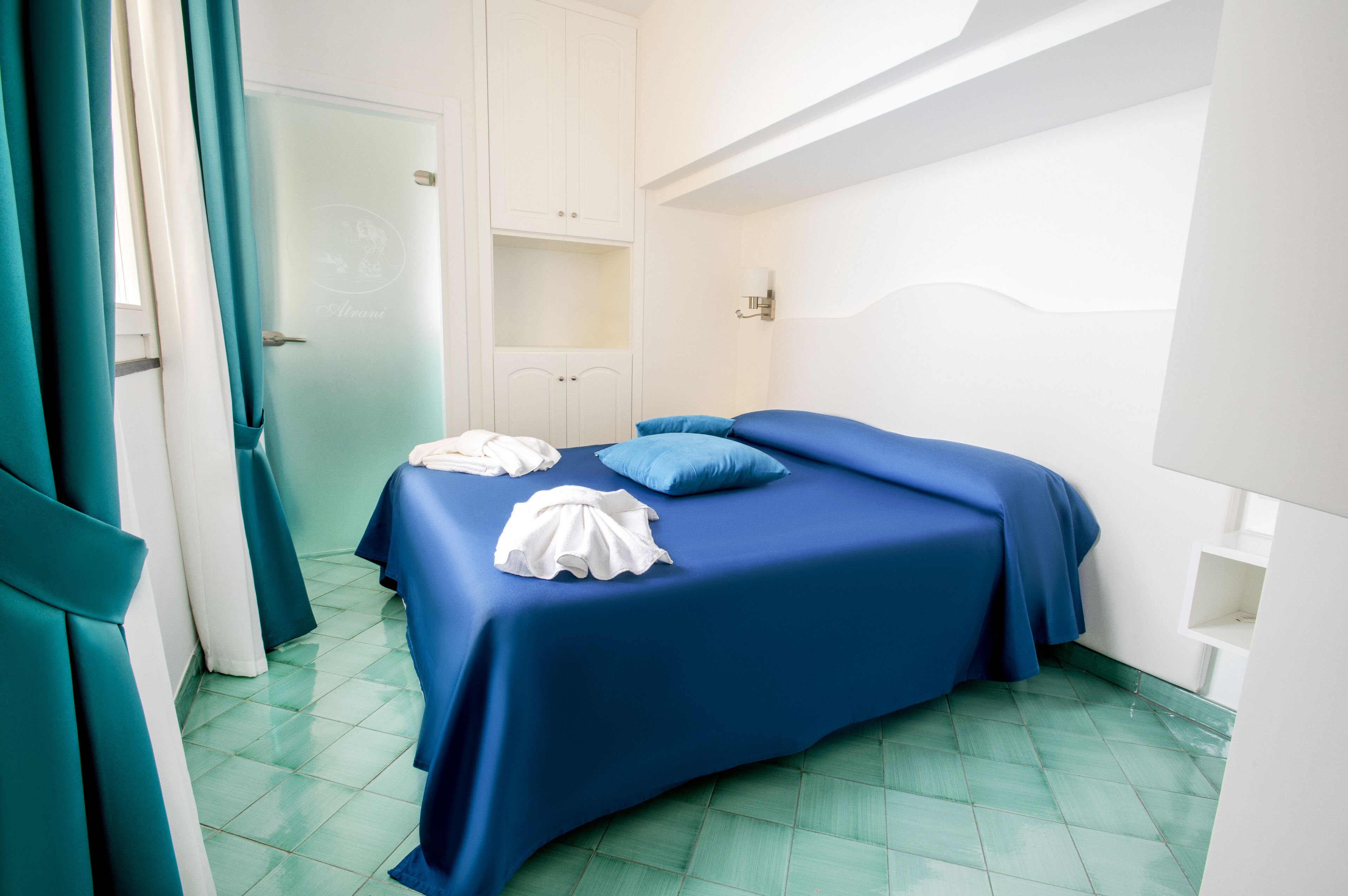 A neatly made bed with blue bedding and white towels, a white wardrobe, and teal curtains.