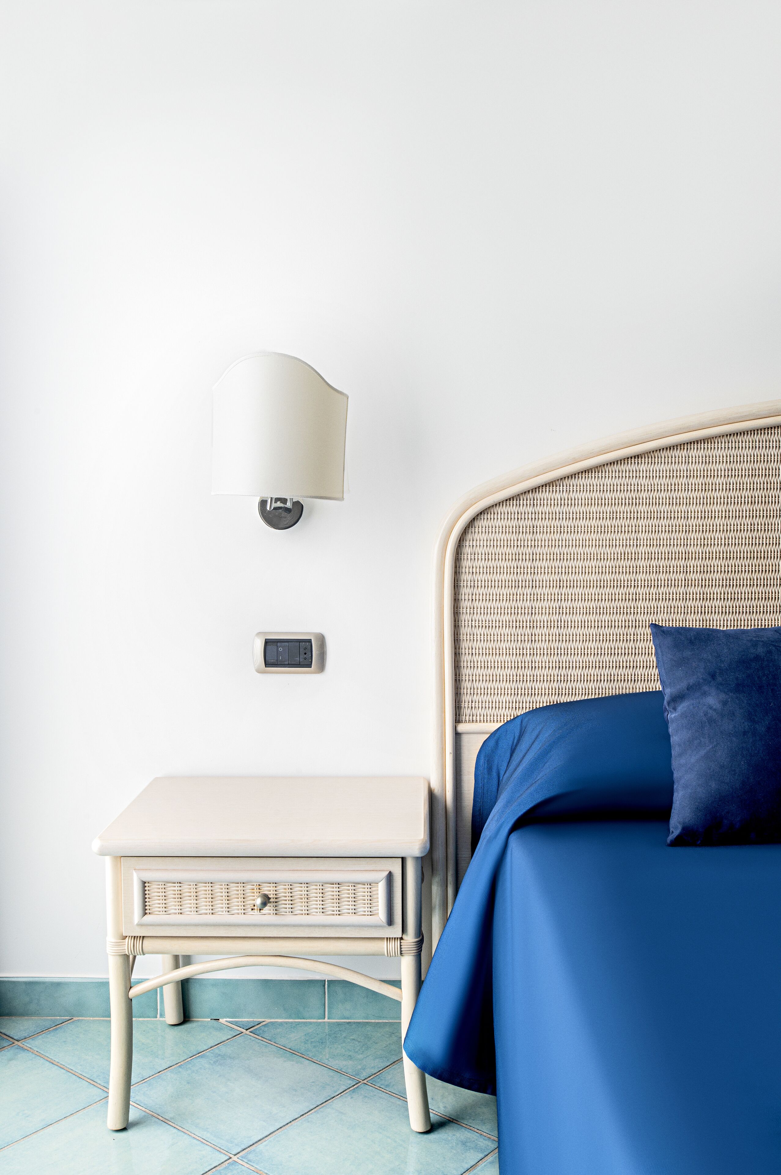 A bed with a woven headboard, a bedside table with a lamp, and a thermostat on the wall.