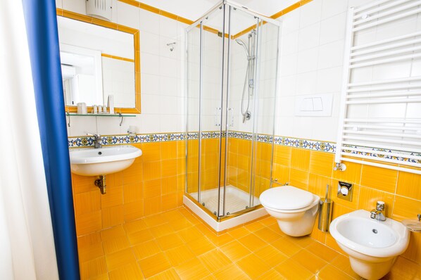 Un baño con azulejos amarillos, ducha con mampara de vidrio, lavamanos, dos inodoros y un bidé.