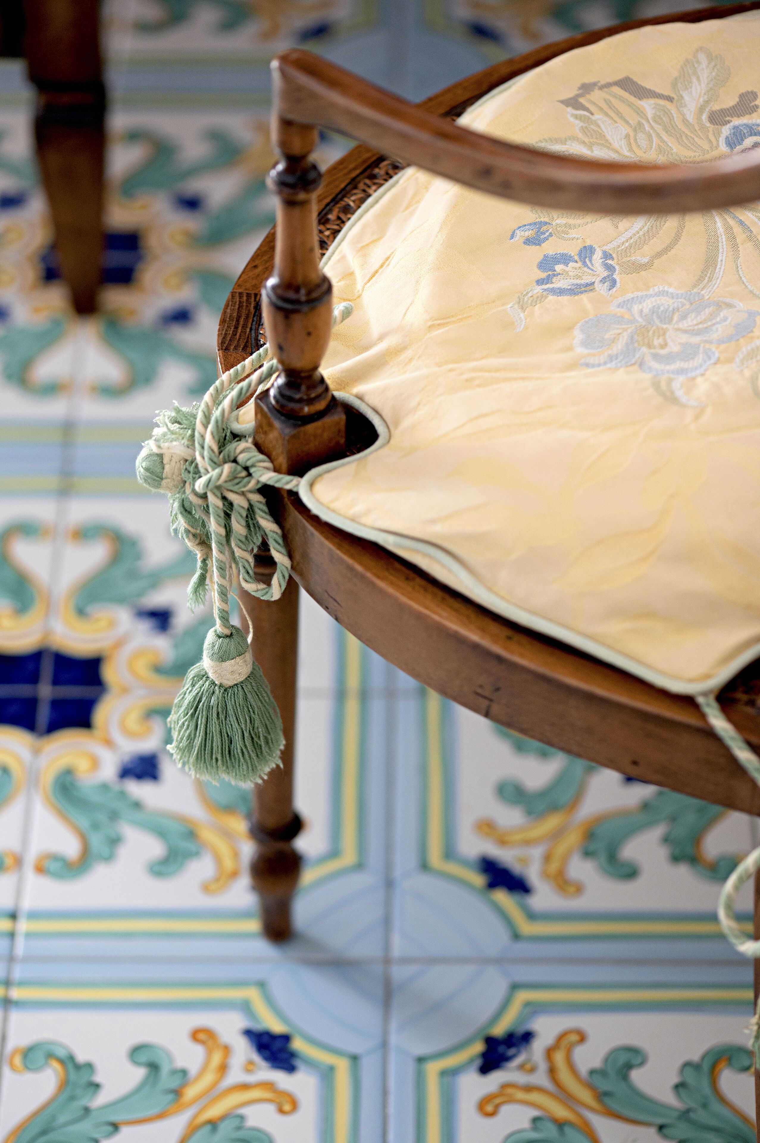 A wooden chair with a cushion and a tassel hanging from the backrest. The floor has a patterned tile design.