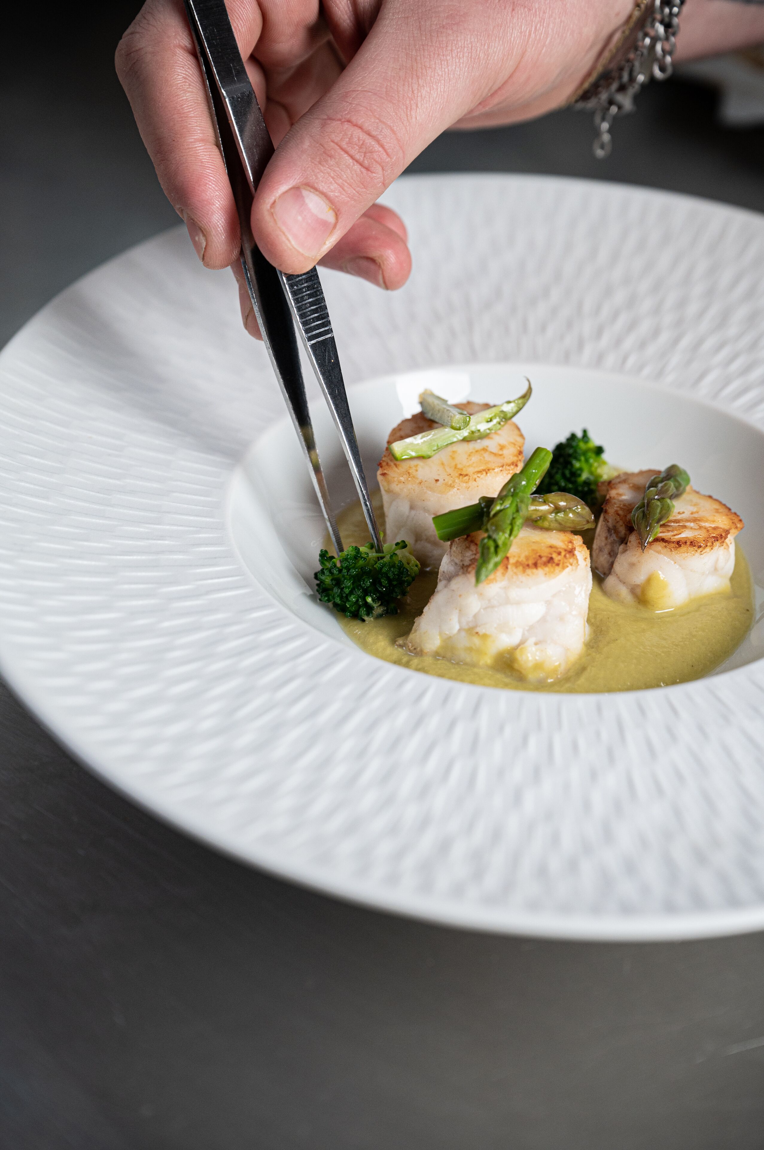A hand using tongs to serve a dish with seared fish and asparagus on a white plate.