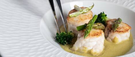 A hand using tongs to serve a dish with seared fish and asparagus on a white plate.