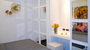 Una habitación con una cama, un armario blanco, una tocador con espejo y una mesita con un jarrón de flores.