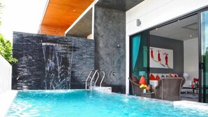 Una piscina moderna al aire libre con cascada, entrada por puerta de cristal y zona de estar con una silla y mesa.