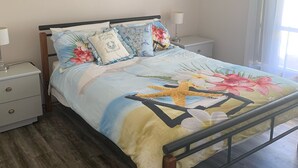 A bedroom with a bed, nightstands, a lamp, an air conditioner, and a painting on the wall. - Sharpie’s Family Shack 🏝 Greenhead.   ☀️ Free Wi-Fi (Green Head)