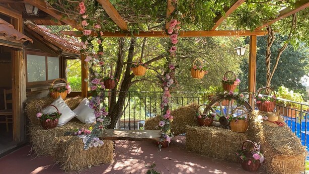 Un espacio de descanso al aire libre con fardos de heno, cestas de mimbre con flores y una pérgola de madera.