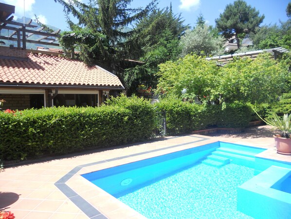 Una piscina amb una terrassa enrajolada, envoltada de vegetació i un edifici amb teulada enrajolada.