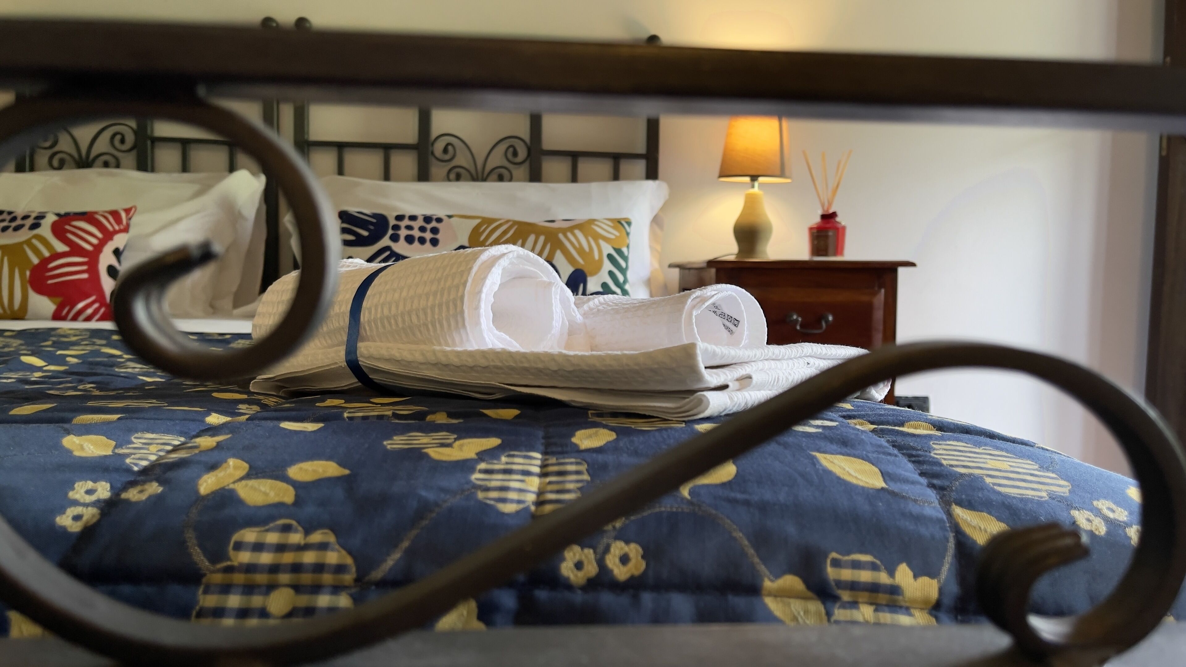 Un lit bien fait, recouvert d’une couvre-lit à motifs floraux bleus, avec une tête de lit en bois et une table de chevet sur laquelle se trouvent une lampe et une bouteille rouge.