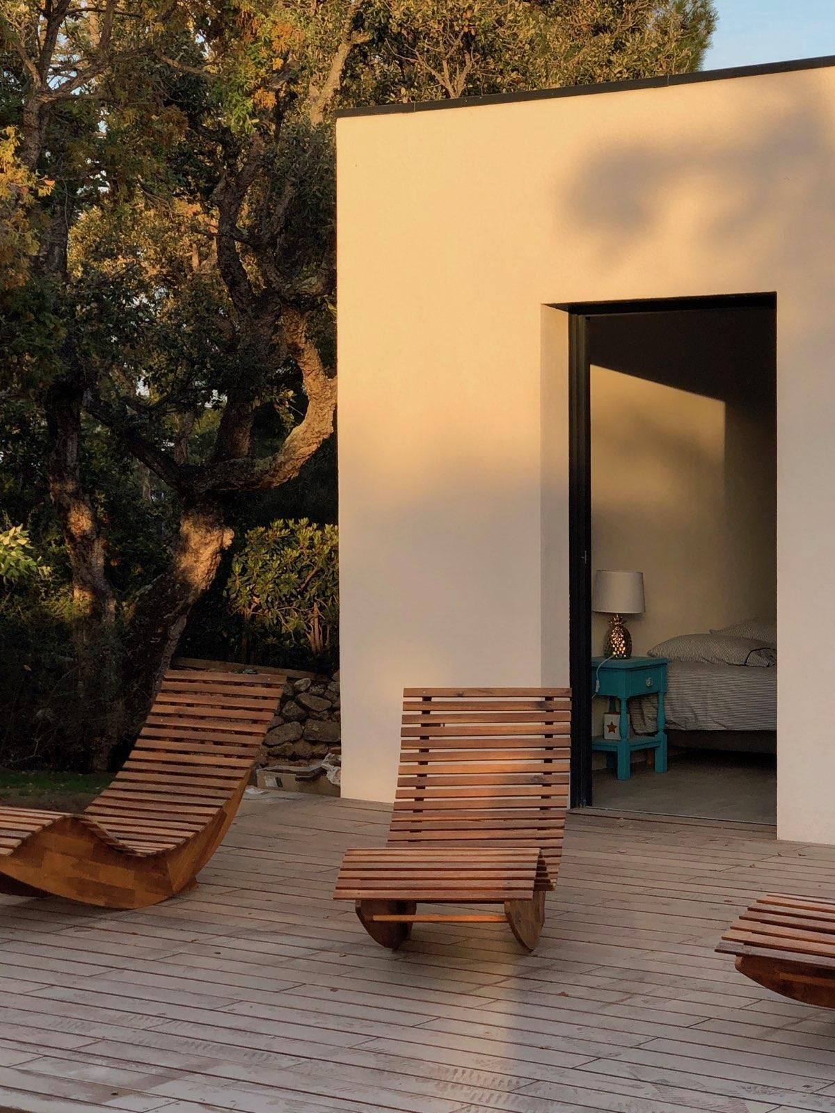 Una terrazza esterna moderna con lettini di legno e vista su una camera da letto attraverso un'apertura.
