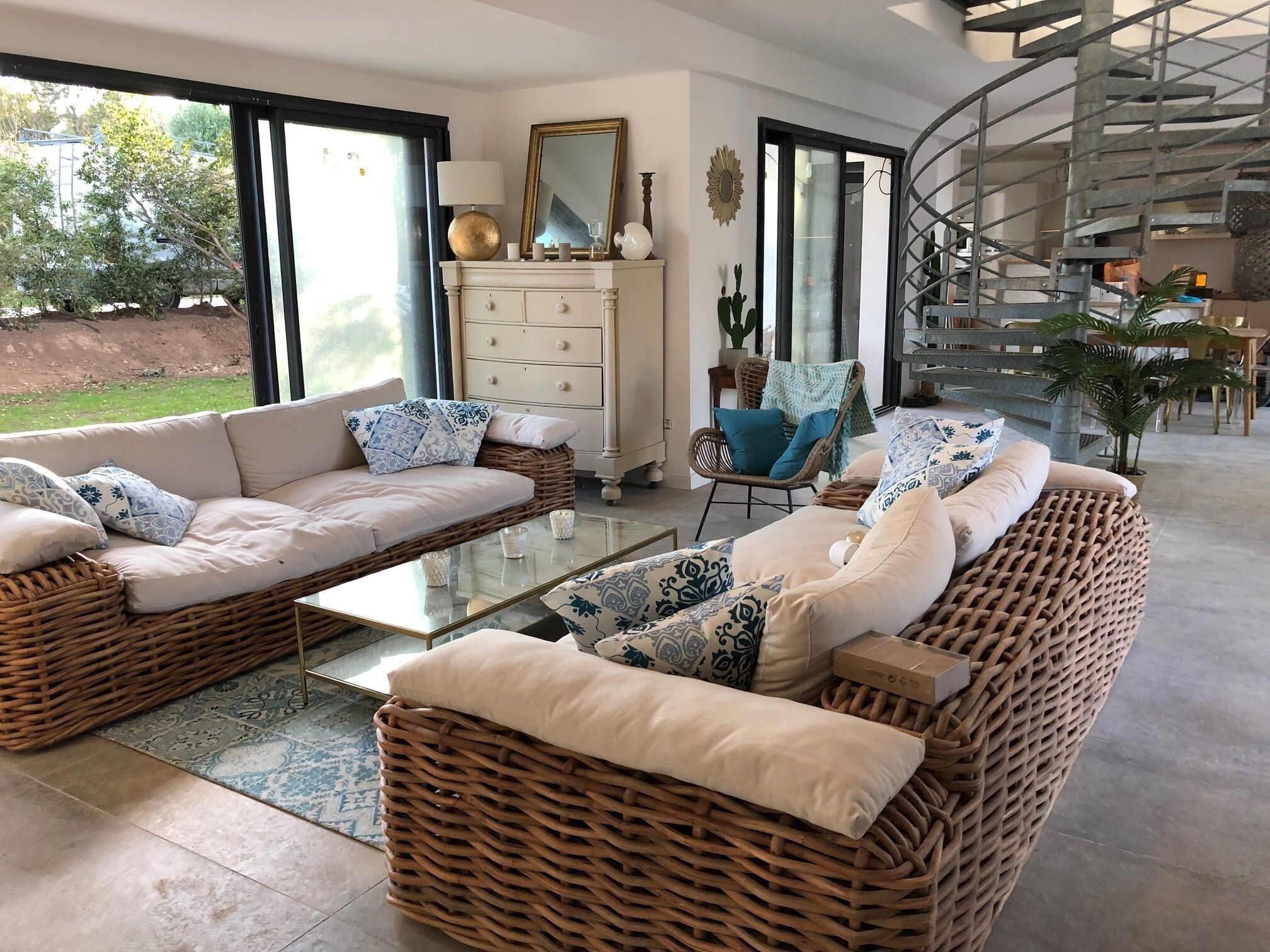 Un vaste salon comprenant un grand canapé, une table basse en verre et des meubles en osier. Un escalier mène à un niveau supérieur, et de grandes fenêtres offrent une vue sur l’extérieur.