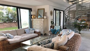 A spacious living room with a large sofa set, a glass coffee table, and wicker furniture. There is a staircase leading to an upper level, and a view of the outdoors through large windows. - South Corsica, Villa 14 p, swimming pool, beach at 300m (Sainte-Lucie de Porto-Vecchio)