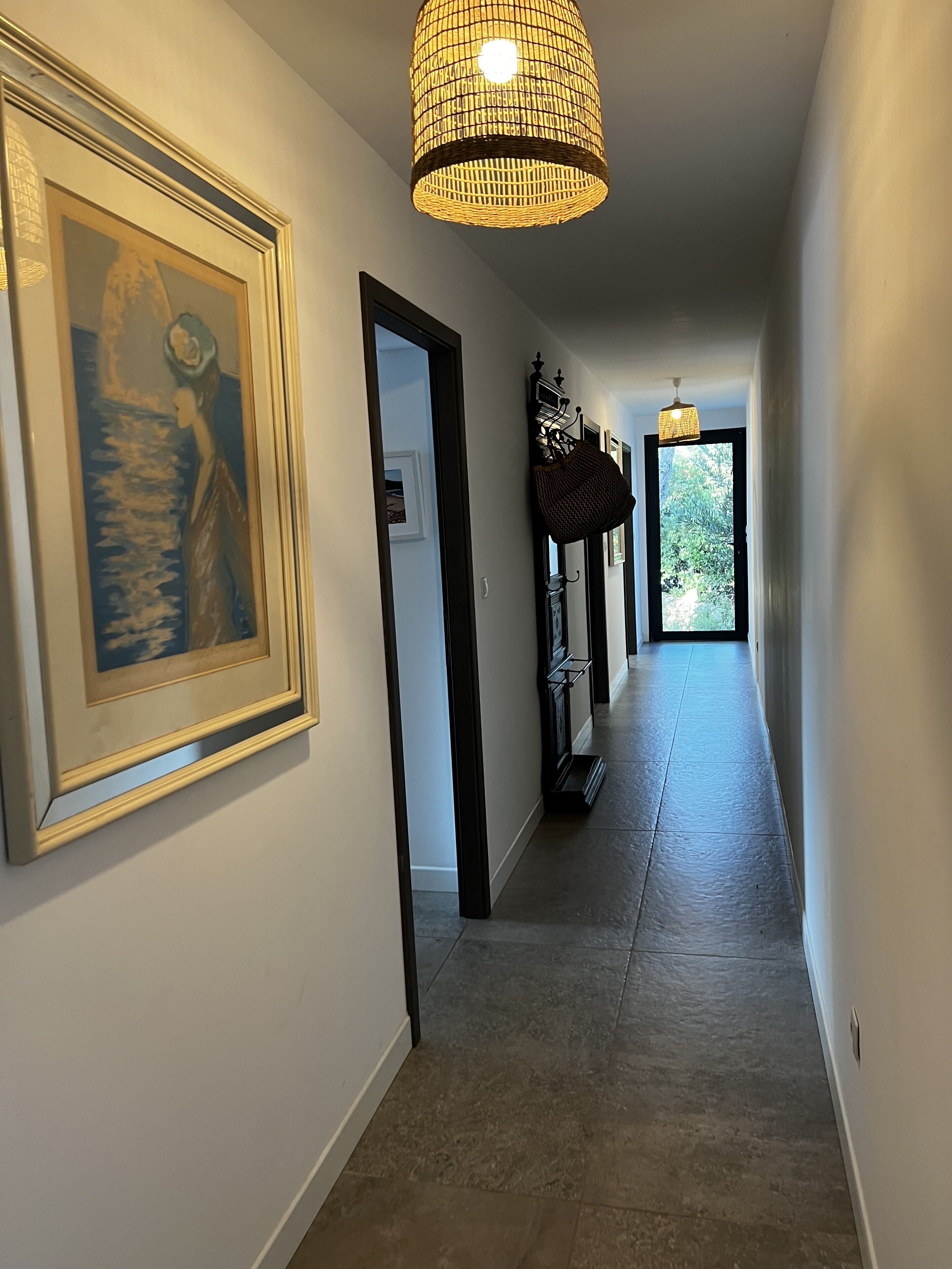 A hallway with framed artwork on the walls, a hanging light fixture, and a view of greenery outside a door.