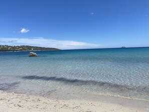 A clear beach with a large rock in the water, a distant island, and a hilly landscape with buildings.