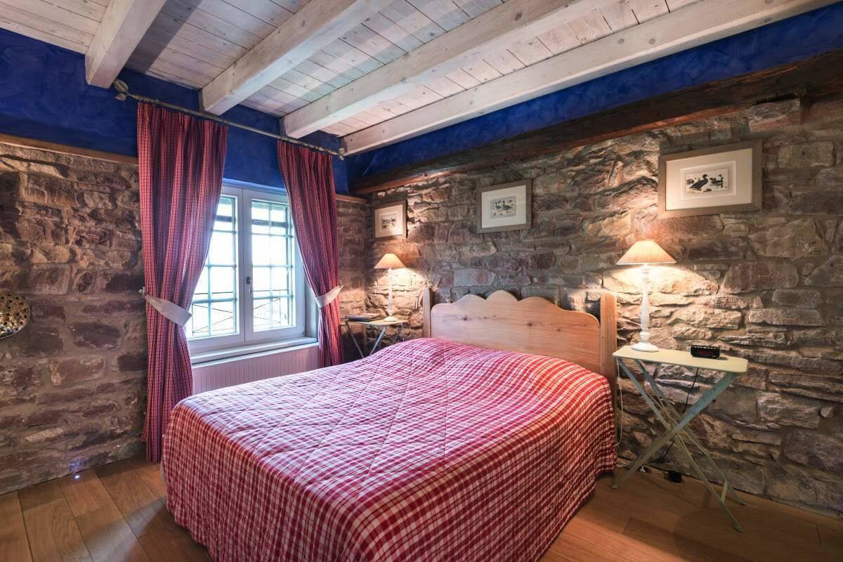 Une chambre avec un mur en pierre, un plafond en bois, un lit recouvert d’une couverture à carreaux rouges, deux lampes de chevet et une fenêtre avec des rideaux rouges.