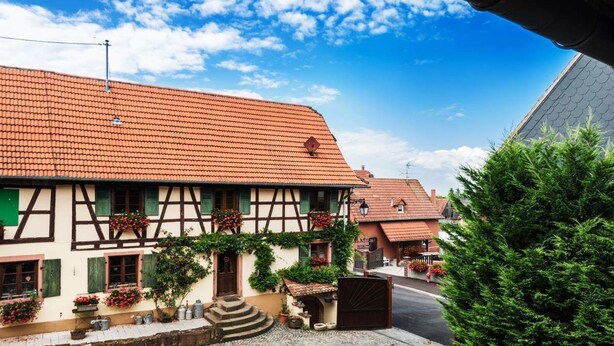Un bâtiment traditionnel à colombages, surmonté d’un toit de tuiles rouges, donnant sur une rue pavée et bordé d’un petit jardin agrémenté de plantes en pot et de fleurs.