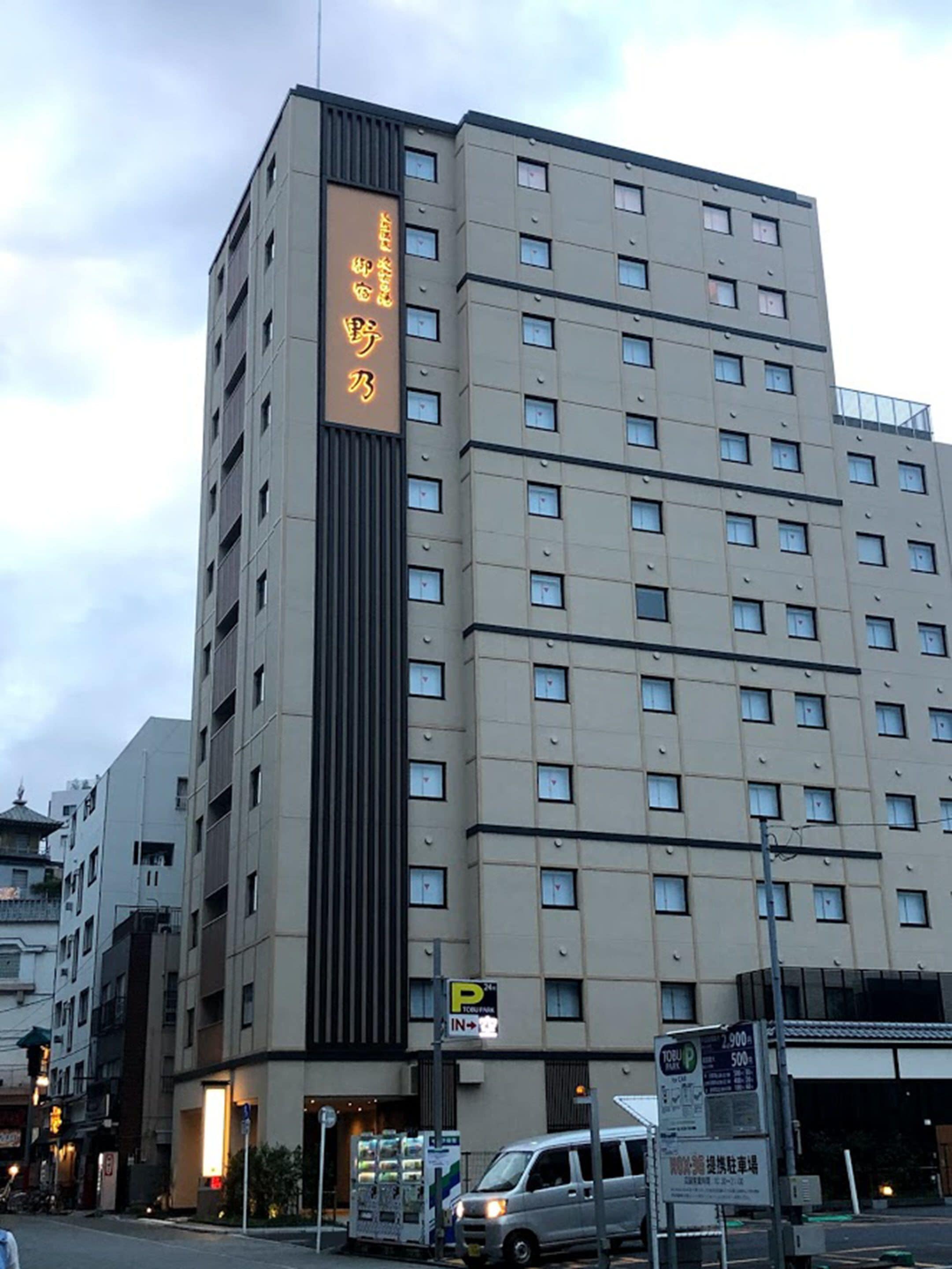 Um hotel com vários andares, com uma placa vertical chamativa na lateral, sinalização de estacionamento e uma máquina de vendas automática na parte externa.
