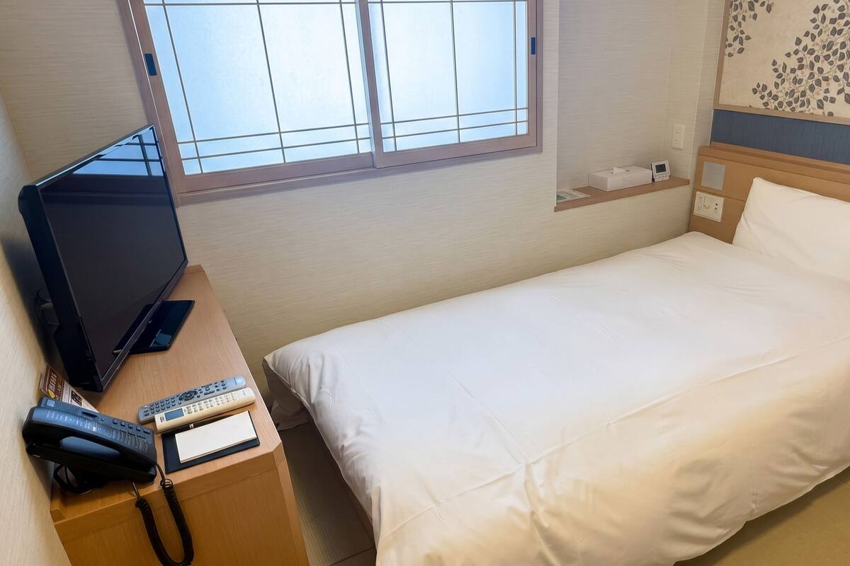 A single bed with white bedding, a bedside table with a telephone, a television, and a small shelf with a remote control.