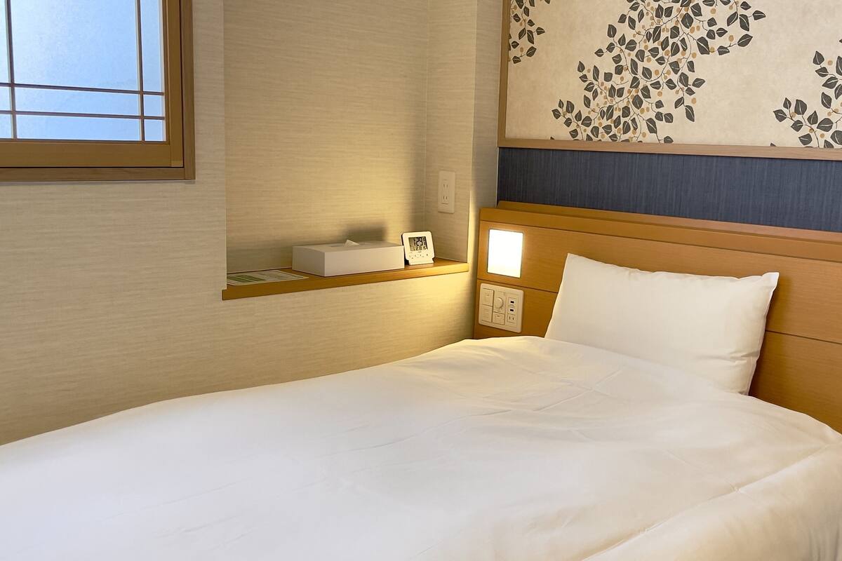 A neatly made bed with white linens, a bedside table with a tissue box and a digital clock, and a wall with a leaf-patterned design.