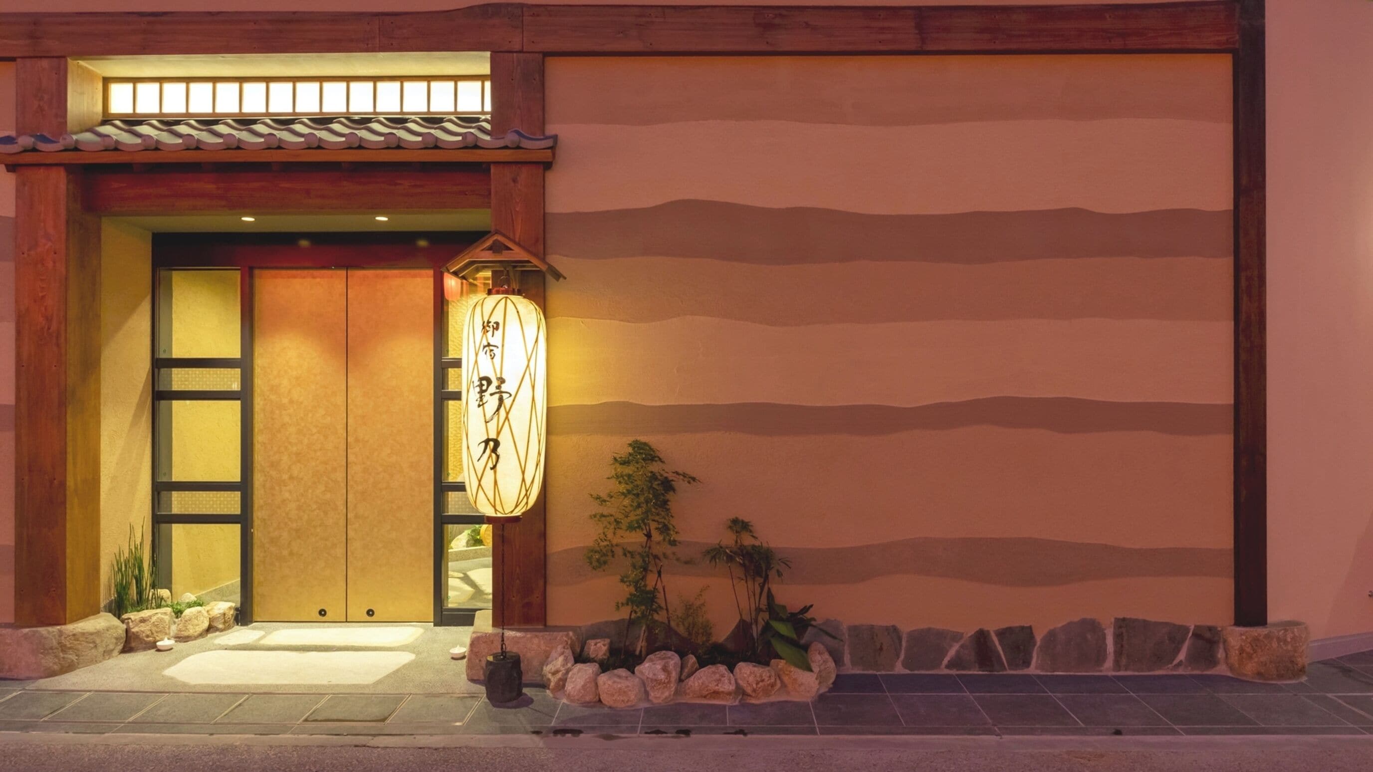 A traditional Japanese building entrance with a sliding door, a lantern, and a landscaped area with rocks and plants.