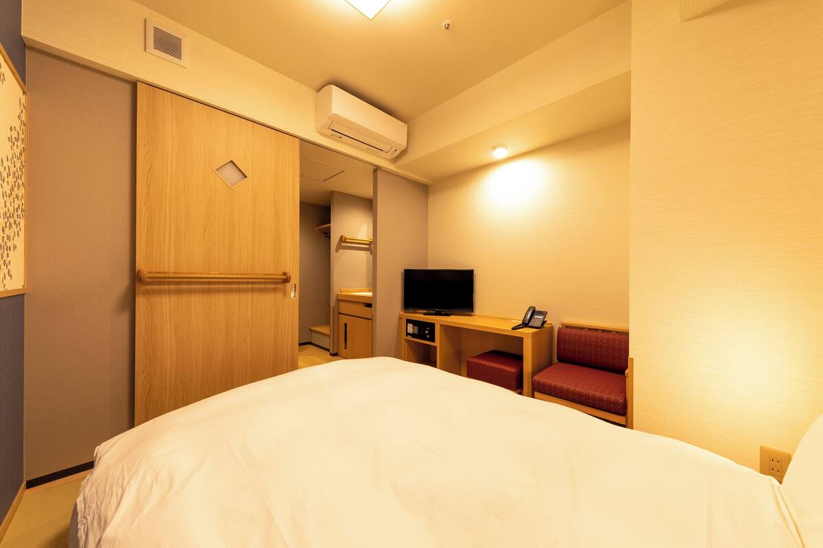 A hotel room with a bed, a desk with a television, a red chair, and a wardrobe.