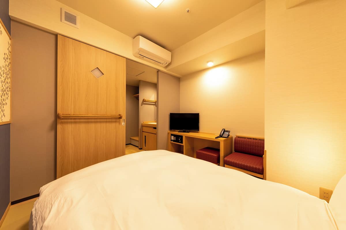 A hotel room with a bed, a desk with a television, a red chair, and a wardrobe.