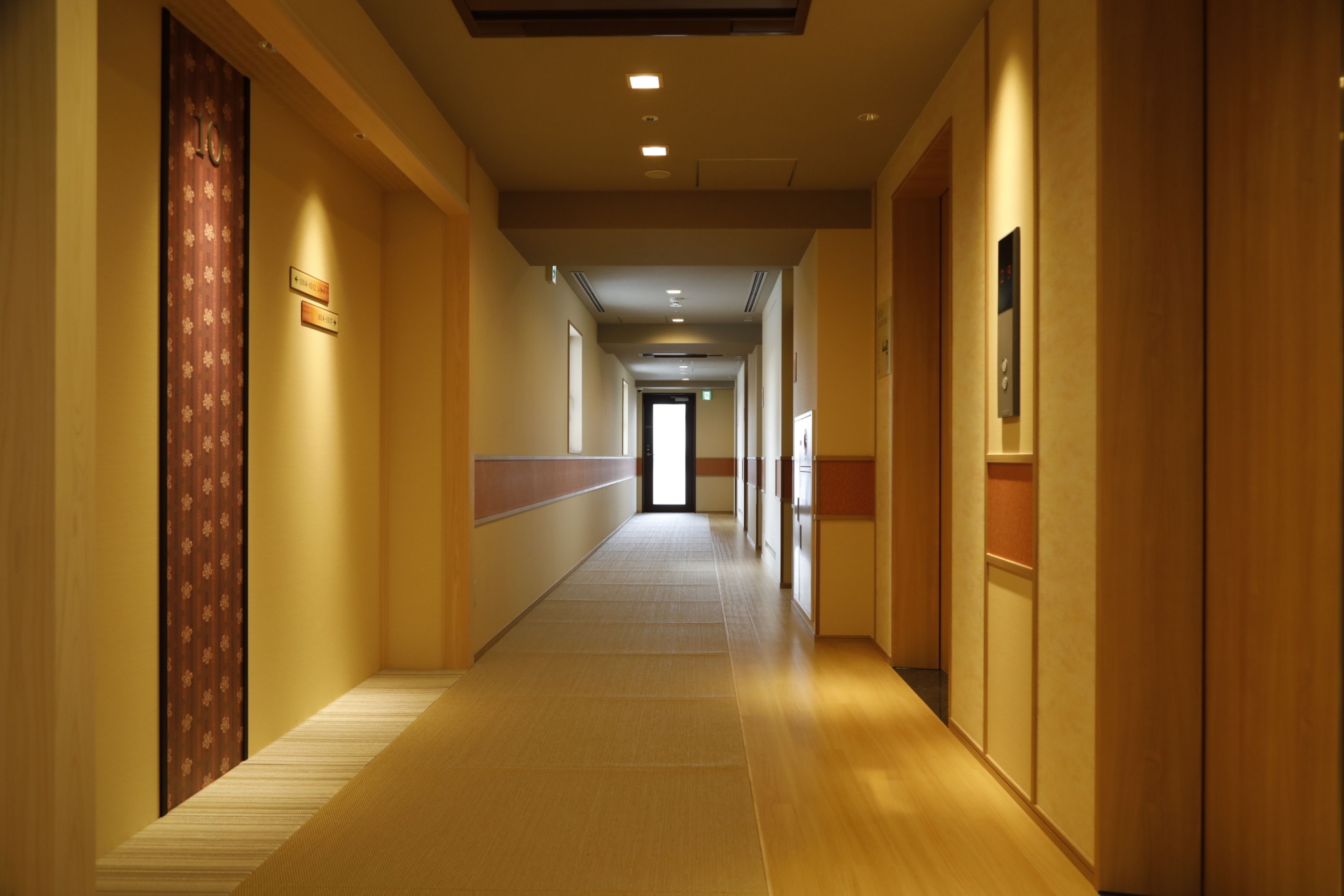 A long, narrow hotel corridor with wooden floors and walls, featuring a door with a floral pattern on the left and a door with a sign on the right.