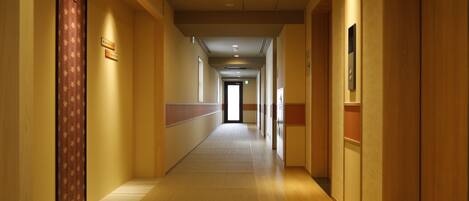 A long, narrow hotel corridor with wooden floors and walls, featuring a door with a floral pattern on the left and a door with a sign on the right.