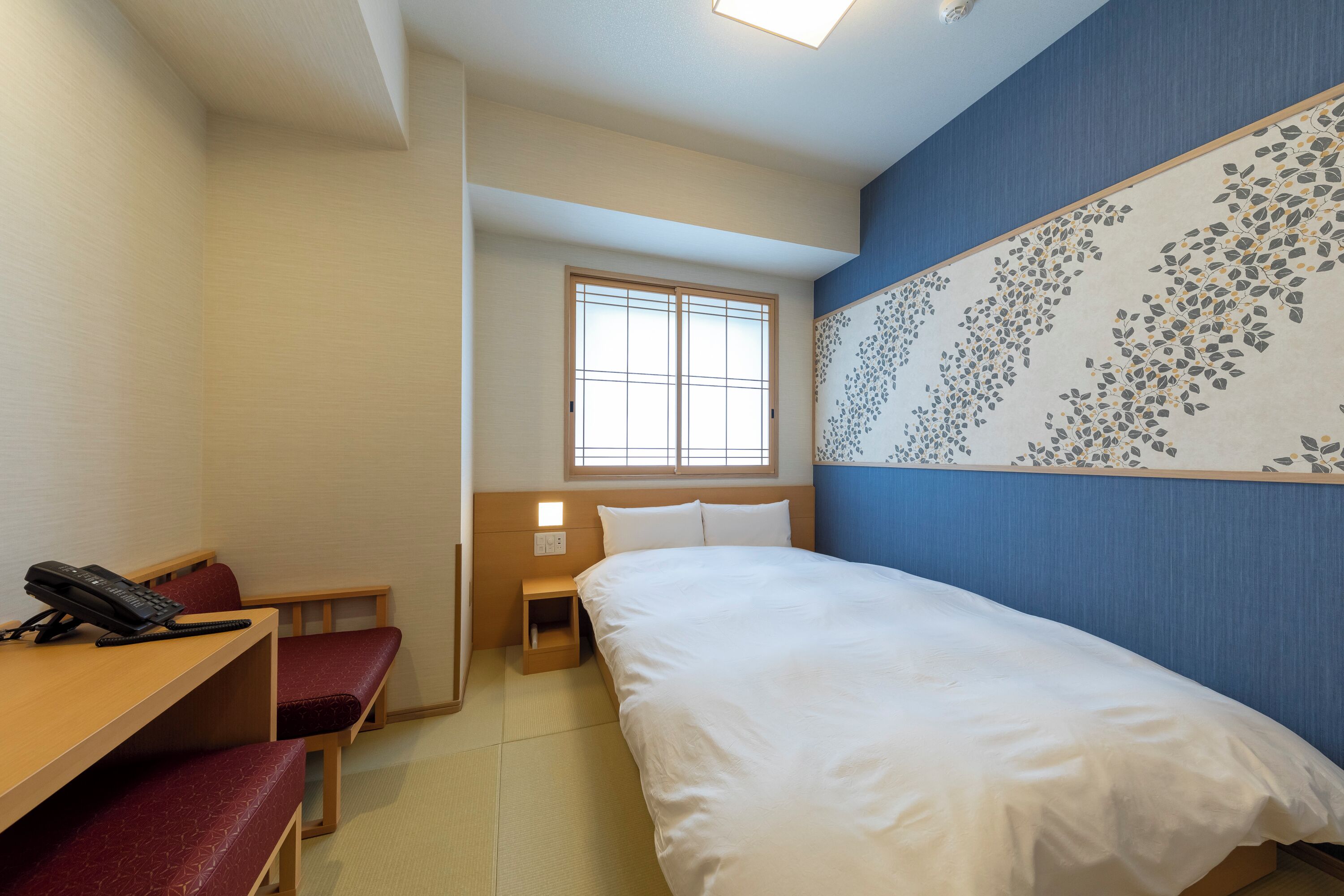 A hotel room with a bed, a desk with a phone, a bench, a small bedside table, and a window.