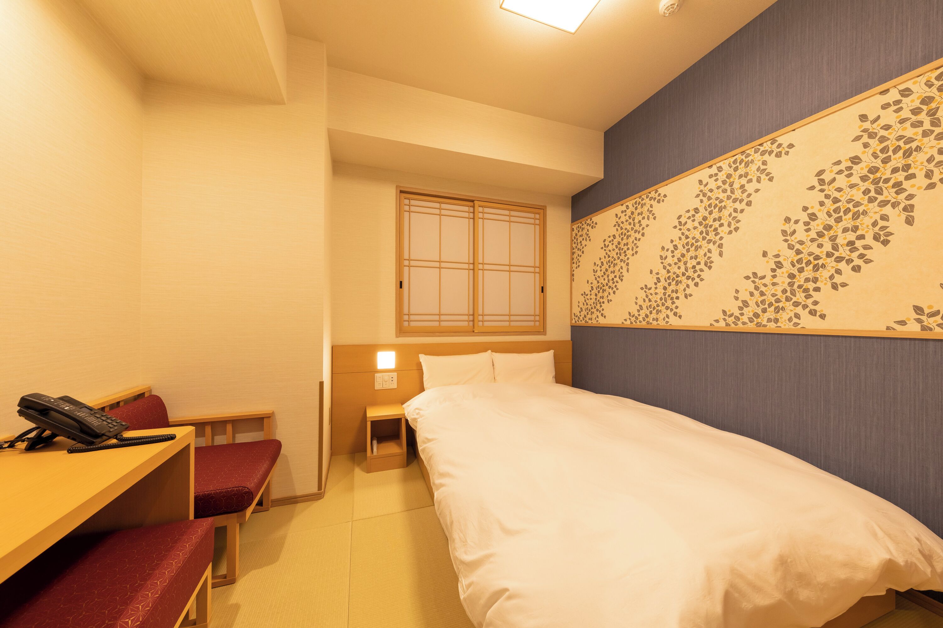 A hotel room with a bed, a desk with a telephone, a bench, a nightstand, and a decorative wall panel.