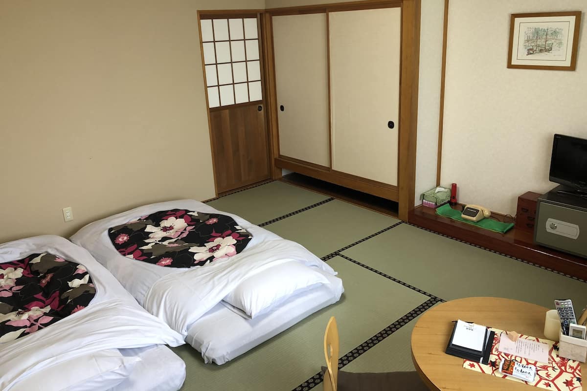 Ein traditioneller japanischer Raum mit Tatami-Böden, zwei Betten mit floralem Muster, einem Schreibtisch mit Computer, einem Fernseher und einem kleinen Tisch mit einer Speisekarte.