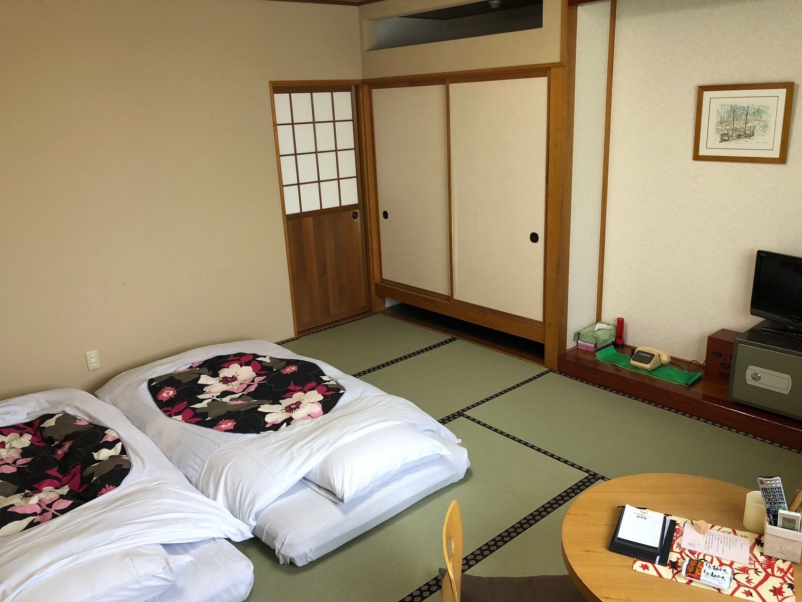 A traditional Japanese room with tatami flooring, two beds with floral bedding, a desk with a computer and phone, and sliding doors.