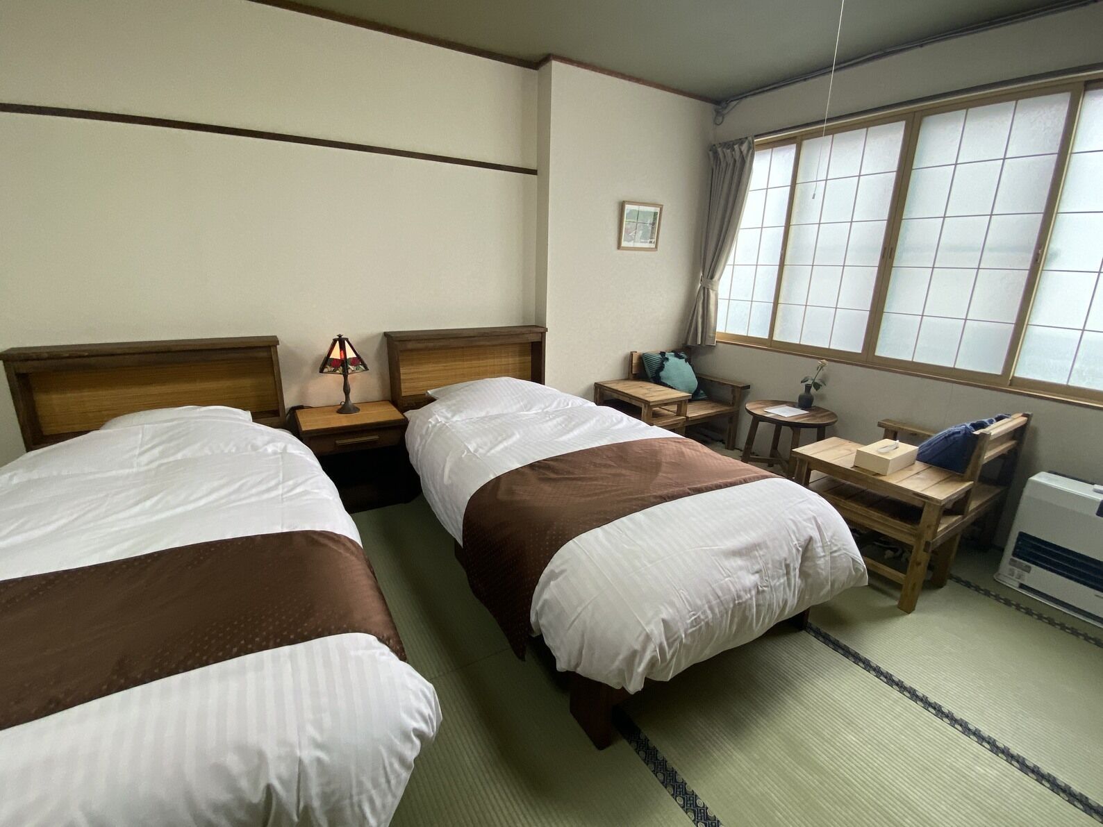A hotel room with two beds, a bedside table with a lamp, a small table with a plant, a window with curtains, and a radiator.
