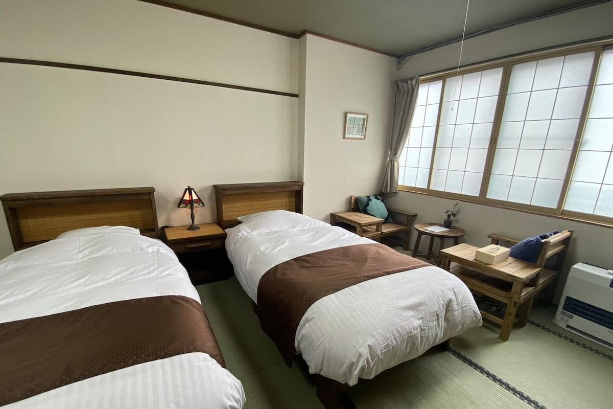 Ein Hotelzimmer mit zwei Betten, einem Nachttisch mit Lampe, einem kleinen Tisch mit Pflanze, einem Fenster mit Vorhängen und einer Heizung.