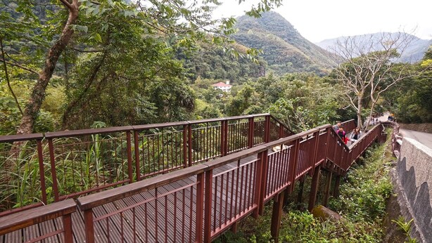 Un pont piétonnier en bois, doté de garde-corps métalliques, entouré d’une végétation luxuriante et avec des montagnes en arrière-plan.