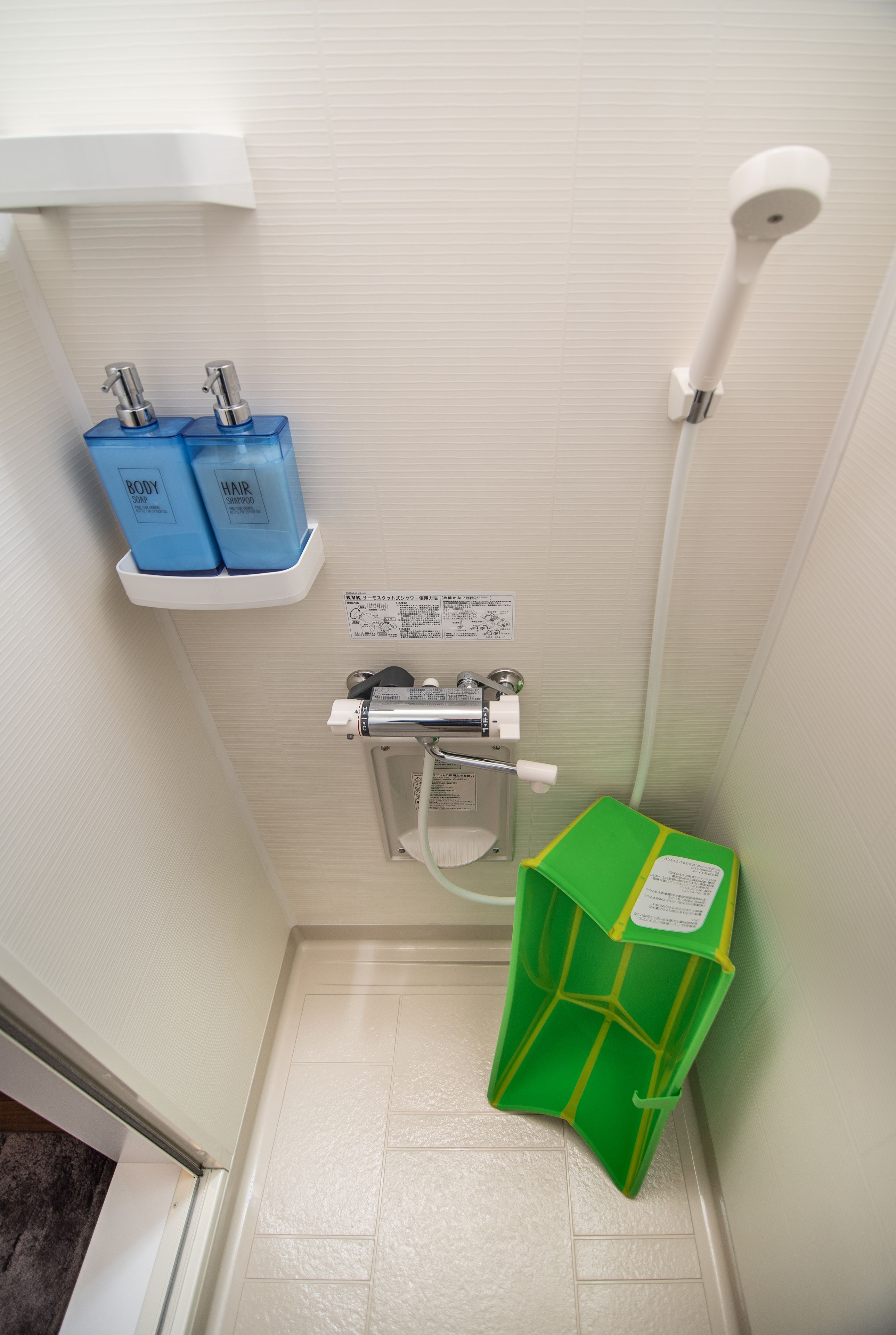 A bathroom with a white sink, two blue bottles labeled "BODY" and "HAIR," a green trash bin, and a white showerhead.