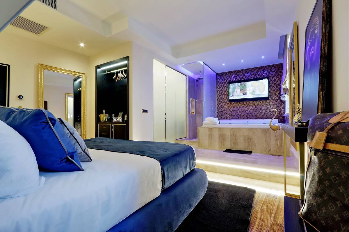 A modern hotel room with a large bed, a bathroom with a bathtub, and a TV mounted on the wall.
