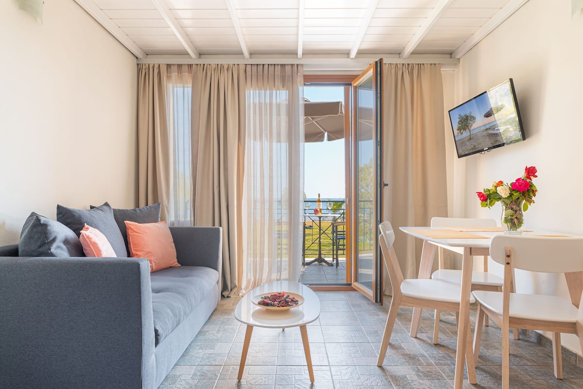 A living room with a sofa, a small table with a bowl of fruit, a TV mounted on the wall, and a balcony with a table and chairs.