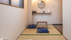 Une pièce avec un sol recouvert de tatamis, un lit avec une literie blanche, une petite table, un miroir et une étagère avec une bouilloire et des tasses.