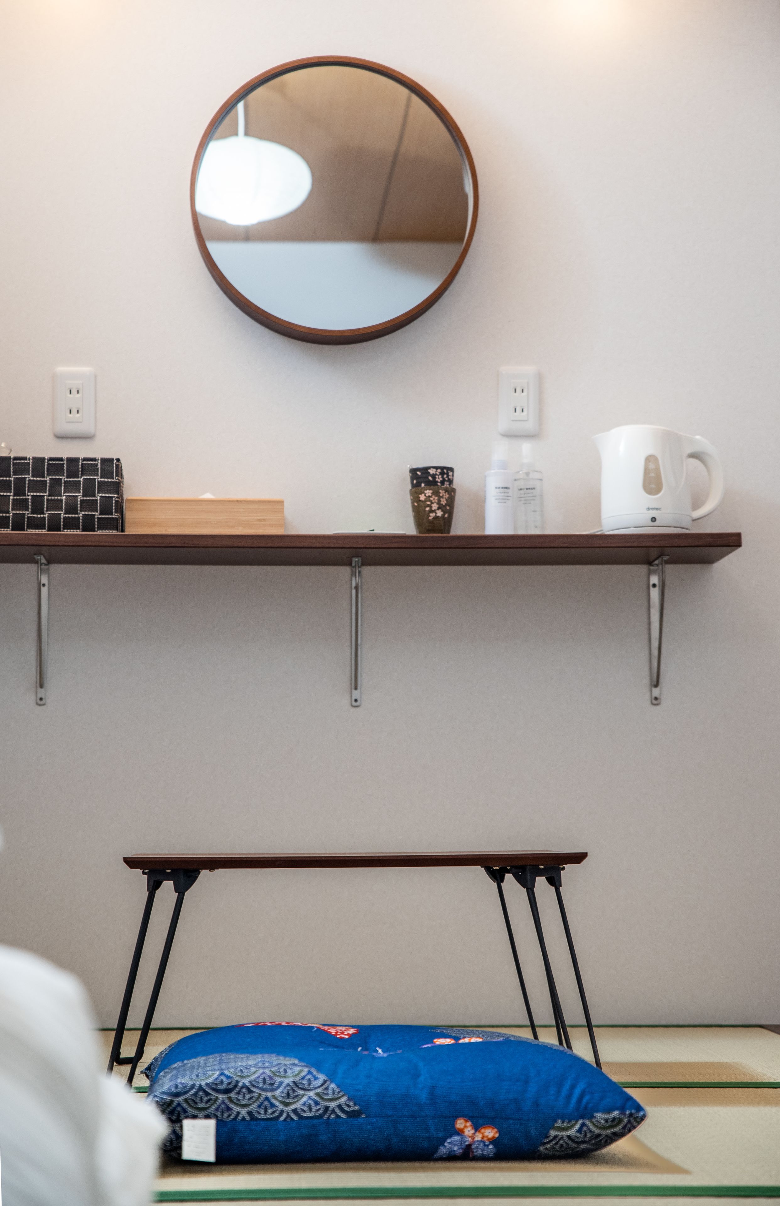 A room with a round mirror on the wall, a shelf with a kettle, a mug, and toiletries. There is a bench with a blue pillow and a patterned cushion.