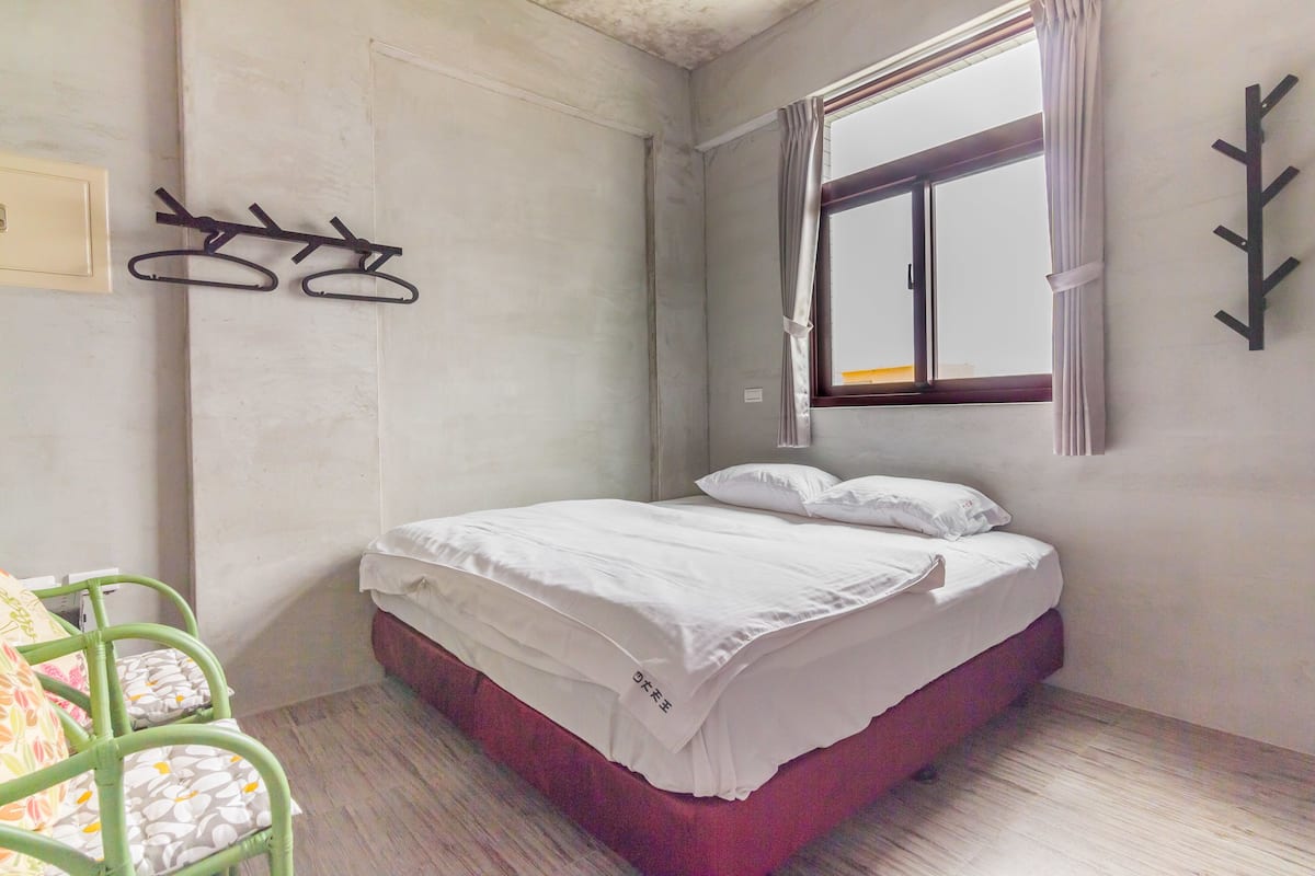 A bedroom with a single bed, a window with curtains, a wall-mounted coat rack, and a green chair with patterned cushions.
