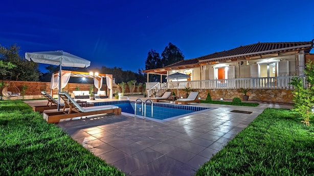 A well-lit outdoor pool area with lounge chairs, a pergola, and a stone house with a balcony. - Villa Eleon with private pool, BBQ and beautiful garden. (Zakynthos)